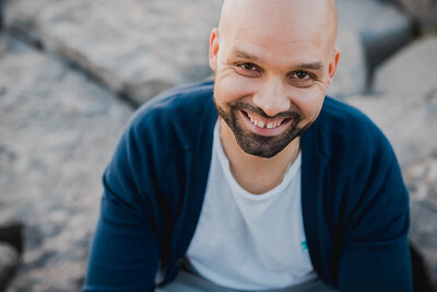 A headshot of a  bald man with dark eyes, moustache and beard wears a white t-shirt under a dark blue cardigan sits on rocks and smiles at the camera