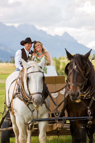 The bride and groom ride together on a horse-drawn carriage, both smiling as they travel through the Jackson Hole countryside, with the Grand Teton mountain range behind them.