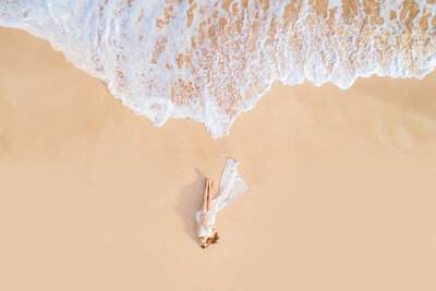 Drone portrait of woman in long white dress laying on the beach during a Maui babymoon photography session