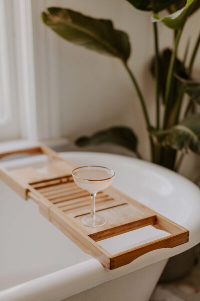 white bathtub with light bamboo wood bathtub tray, cocktail glass with pink cocktail and gold rim, potted plant blurred in background