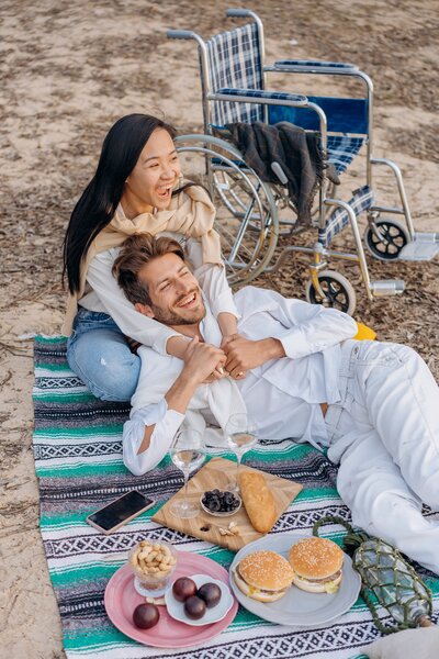 Two partners of differing gender presentations rest on a picnic blanket. They are holding hands, while one sits cross-legged, and the other lies in their laptop. They are smiling past the camera, with a sandy beach and wheelchair visible in the background.