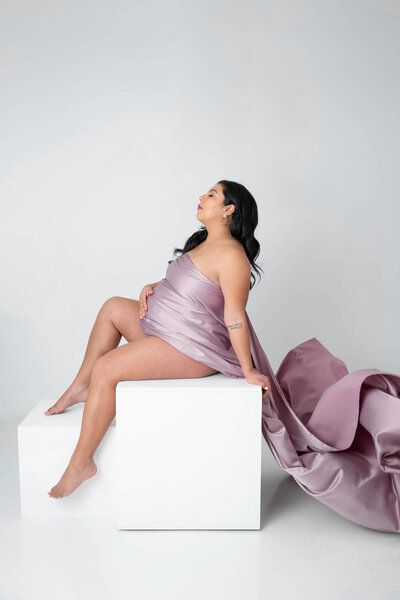 A beautiful mother draped in silky mauve fabric. posing on a white box.