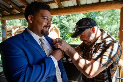 Groom in a blue suit smiles as his elopement photographer pins a boutonniere on his lapel at an outdoor wedding venue.