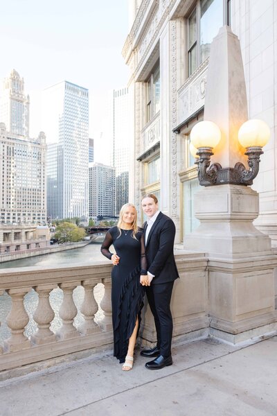 An elegantly dressed couple poses on the Chicago Riverwalk near the Wrigley Building, with the river and high-rise cityscape in the background. The woman wears a black dress with sheer sleeves and a ruffled slit, while the man wears a black suit and white shirt.








