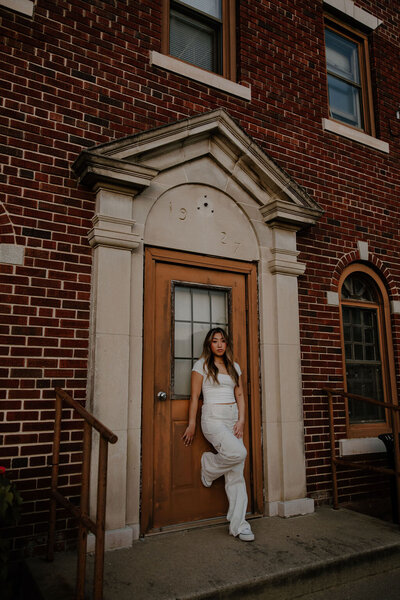Senior picture of a girl standing, leaning against a beautiful old brick building in white pants, shoes and short sleeved shirt