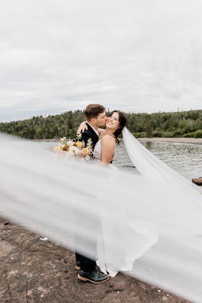 Bride and groom during an intimate elopement in Grand Marais, MN, captured by a wedding and elopement photographer in Duluth, MN. The bride’s veil is pulled slightly in front of the camera for a romantic effect as she pops her foot up while her husband kisses her cheek, with a scenic North Shore backdrop.