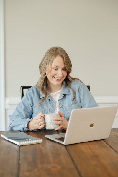 Woman wearing a light blue jean jacket holding a cup of coffee at a wooden table smiling at a Macbook Pro