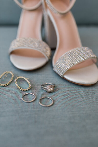 A close-up of bridal accessories, including embellished high heels, wedding rings, earrings, and a diamond engagement ring, arranged on a soft blue surface