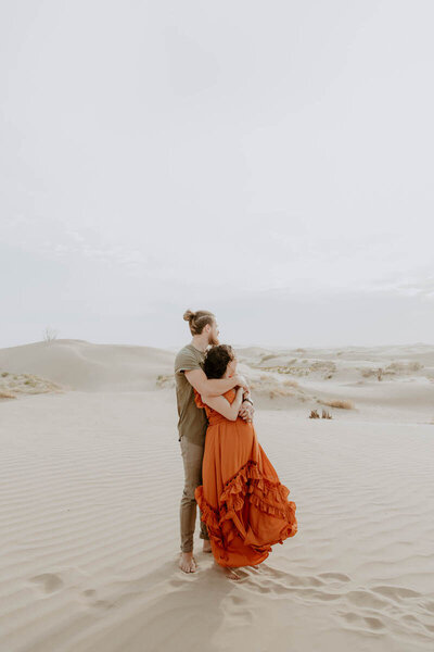 Boho couple wearing neutral color outfits holding each other looking off into the distance at the Sand Dunes