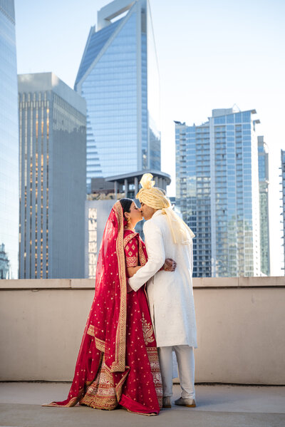 Indian wedding couple kissing on the rooftop of the Grand Bohemian Hotel Charlotte, with the city skyline in the background. Bride in traditional red lehenga and groom in ivory sherwani with turban. Captured by DeLong Photography, Charlotte Indian wedding photographers specializing in vibrant cultural wedding photography