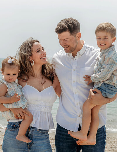 Beach family photography by Stamford CT photographer showing parents with two young children in casual white and striped outfits with ocean backdrop