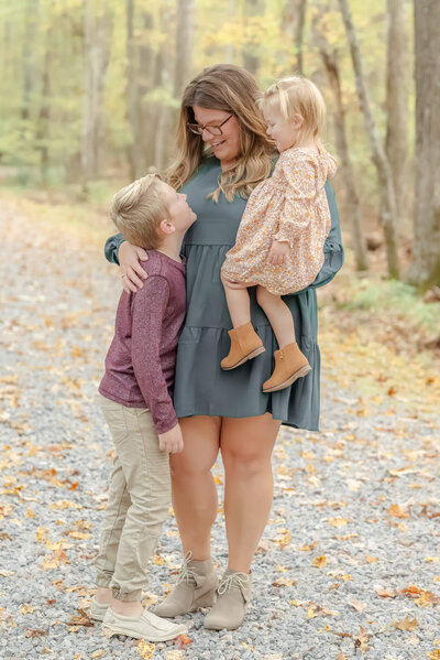 A mother, wearing a teal dress, holds her toddler daughter and hugs her son. They are having fall photos done by their Chesapeake photographer.