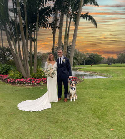 a bride and groom next to each other with a australian shepherd (grey, white and tan fluffy dog) beside the groom sitting down. Dog is wearing a wedding tux with a beautiful red sky.