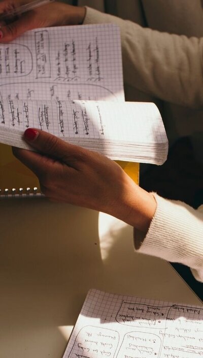 Kaya flipping through handwritten notes in a grid notebook with visible crossed-out lists.