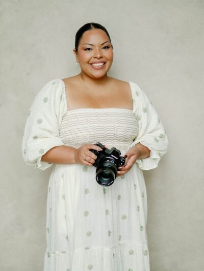 Wedding photographer Jennifer Nolan smiles while holding her camera and wearing a cream dress with green flowers from Anthropologie