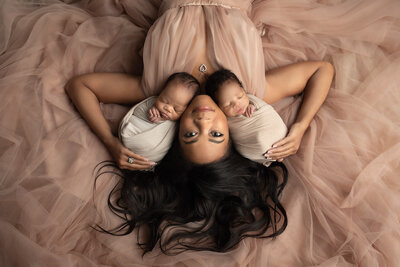 Mother lying in a flowing blush gown holding her newborn twin babies swaddled in neutral wraps, captured in a Dallas photography studio.