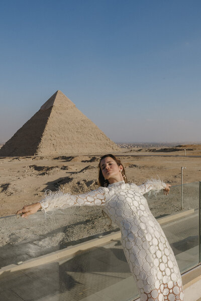 A photo of a model who is wearing bridal editorial white dress and is leaning against the glass rail with the Giza Pyramid in the background. This was taken by Neha.