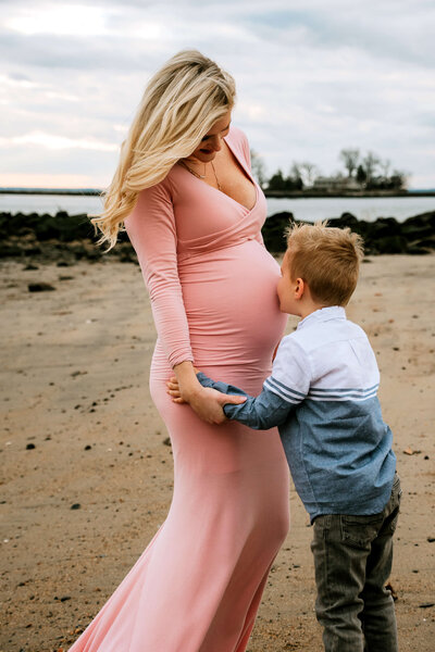 Connecticut maternity photographer captures pregnant mother in pink dress with young son on beach at sunset