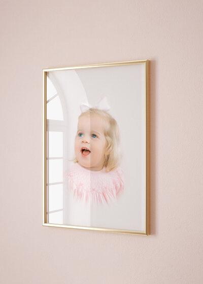 A blonde toddler's heirloom portrait is displayed in a classic gold frame on a pink wall.  Artwork photographed and designed by Charlotte Photographer,  Melissa Mayrie Photography