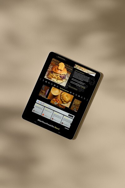 A tablet displaying the website for The Patty Freaks, featuring a large image of a gourmet burger with text describing the dish. The site includes sections showcasing various food items, customer reviews, and the hashtag "#GetYourFreakOnHere!" The tablet is placed on a beige surface with shadows