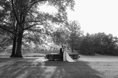 A couple stands beside a classic car under a large tree in a grassy field. The scene is captured in black and white.