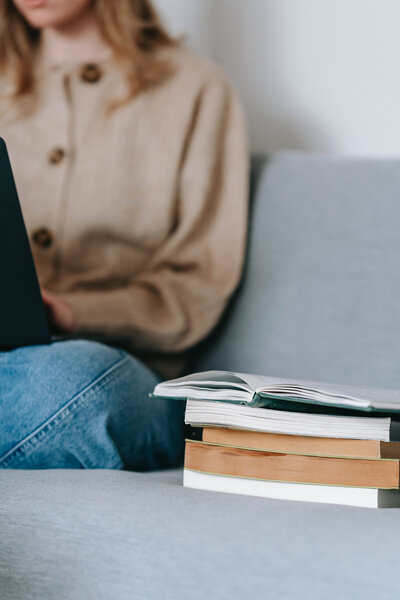 This image shows a woman wearing jeans and a brown sweater sitting on a couch, next to a stack of books, with the topmost book opened to a middle page. She also has a laptop in her lap, and the image is cropped so her face cannot be seen.