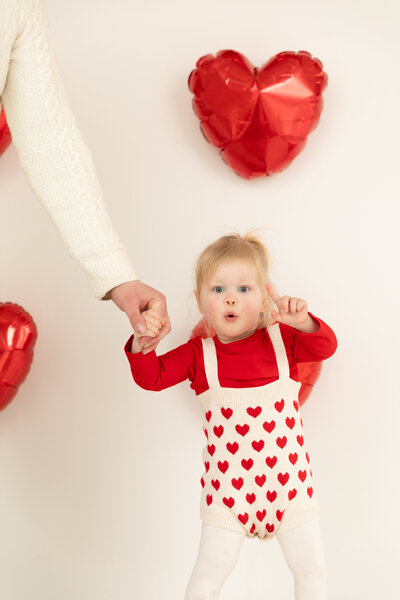toddler girl points to camera for valentine's photo session with red heart balloons on the wall behind her at studiostudio in ann arbor michigan