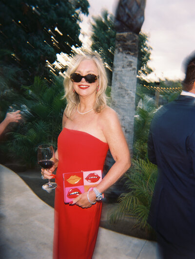 Film photo of blonde woman wearing a red strapless dress and sunglasses holding a clutch and wine glass