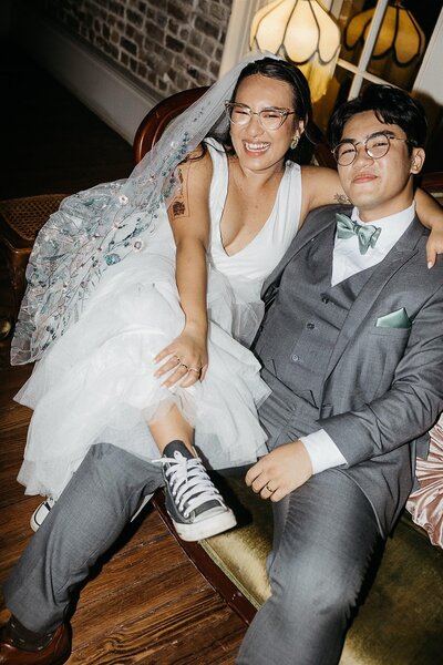 A bride and groom sitting on a couch, the bride wearing Converse sneakers with her wedding dress and laughing, while the groom smiles beside her.