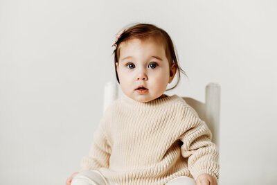 six month old baby girl in a knit sweater sits on a wooden toddler chair during her six month old mini milestone session in Denver.