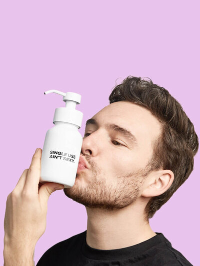 Studio shot of Josh kissing a Single Use Aint Sexy refillable soap bottle against pink background.