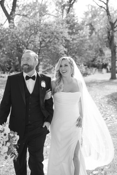 black and white photo of  a bride and groom walking arm in arm smiling with trees and foliage in the background