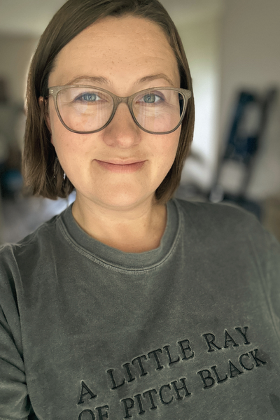 This photo is Kasey, who is a person with short brown hair and glasses smiling at the camera, wearing a gray shirt with text on the front that reads "A little ray of pitch black"