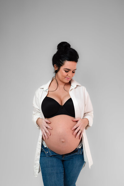 A woman in jeans and a white shirt poses with her belly exposed for her maternity pictures in the sutdio