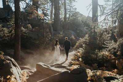 Bride and groom walking away down a dirt path holding hands and looking at each other there is dust catching in the sunlight from their feet on the trail and they are surrounded by large rocks and trees