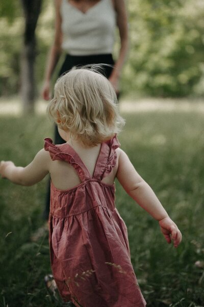 Liten tjej i rosa klänning springer i gräset mot sin mamma.