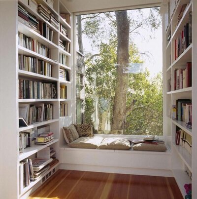 Reading nook under a large window, surrounded by tall white bookshelves full of books. Photo by Safdie Rabines.