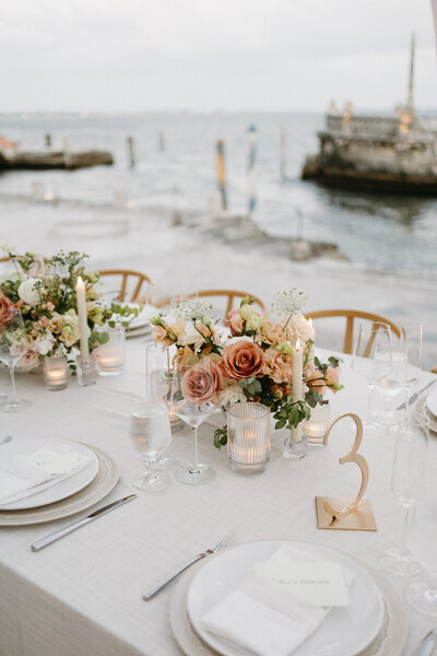 A peach floral centerpiece with gold Table Number 3 on top of a neutral table cloth. Plates and chargers flank each side