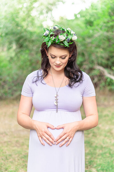 A pregnant woman wearing a lavender dress and a flower crown stands in a small grove of trees at highland glenn park in the summer
