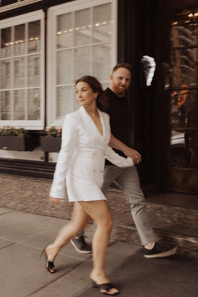 A couple walking briskly past a windowed storefront, the woman in a white blazer dress.