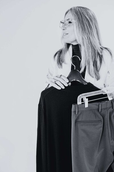 black and white image of a woman holding a black shirt and gray pants on hangers during a styling session