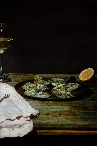 A moody, atmospheric image of oysters on a plate with a halved lemon, set on a rustic wooden table. Photographed by Mica McCook, this still life captures the freshness and elegance of seafood, perfect for a refined dining experience.
