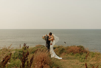 bride and groom share a kiss in front of ocean on wedding day in pei