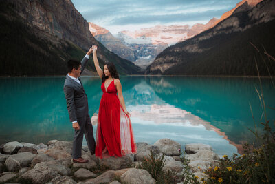A man dressed in a suit goes to twirl his fiancée as they stand in front of a lake with mountains in the background.