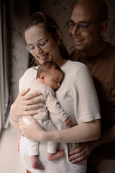 Moeder heeft baby tegen haar borst aan en vader staat erachter. Ze kijken lachend naar de baby