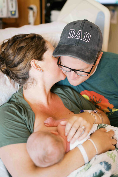Image of husband and wife sitting on hospital bed wife kissing husband's cheek who is wearing a hat saying "Dad" and holding a newborn by Portland Family Photographer Emilie Phillipson Photography