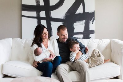 A family of four with a newborn and a toddler sitting on a white couch in front of an abstract painting, captured by a Pittsburgh photographer.