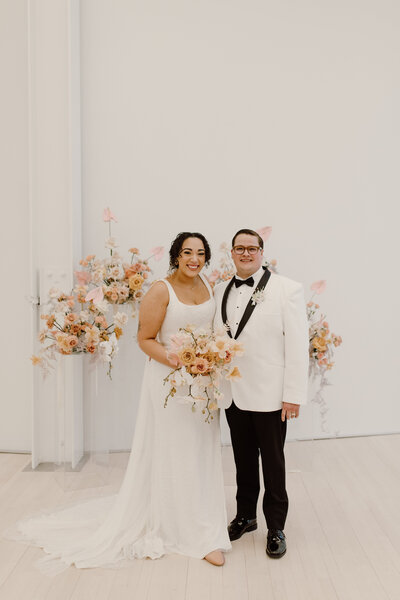 A couple stands for a portrait at their wedding, holding an Anthousai bouquet and surrounded by a large floral installation.