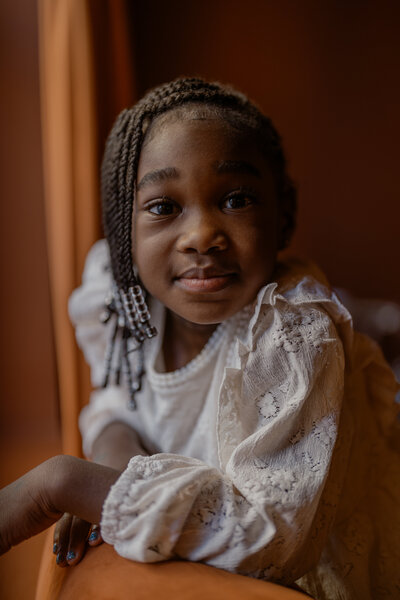 A soulful studio portrait of a young girl wearing an off-white lace dress, smiling warmly into the camera. Captured in natural light against a rich orange backdrop, this image represents a childhood milestone session for ages 5 to 12.