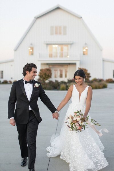 Bride and groom smile and walk holding hands in front of barn wedding venue